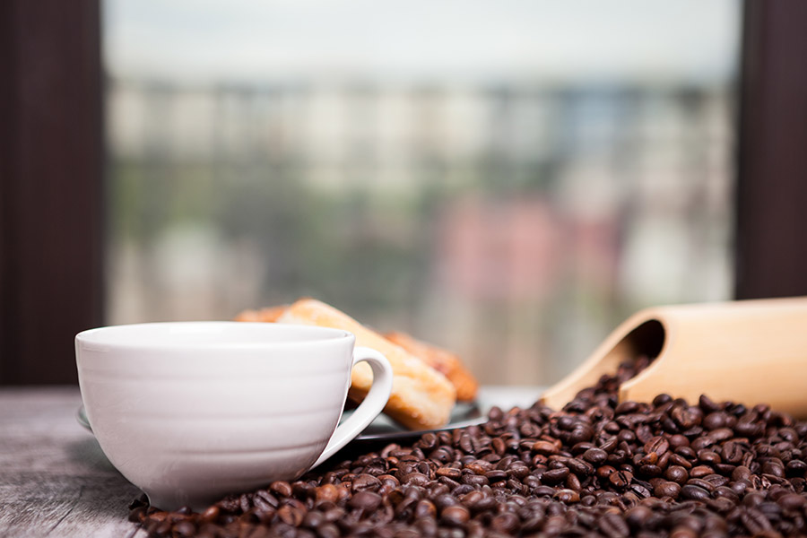 Weiße Kaffeetasse neben verstreuten Kaffeebohnen und Gebäck im Hintergrund